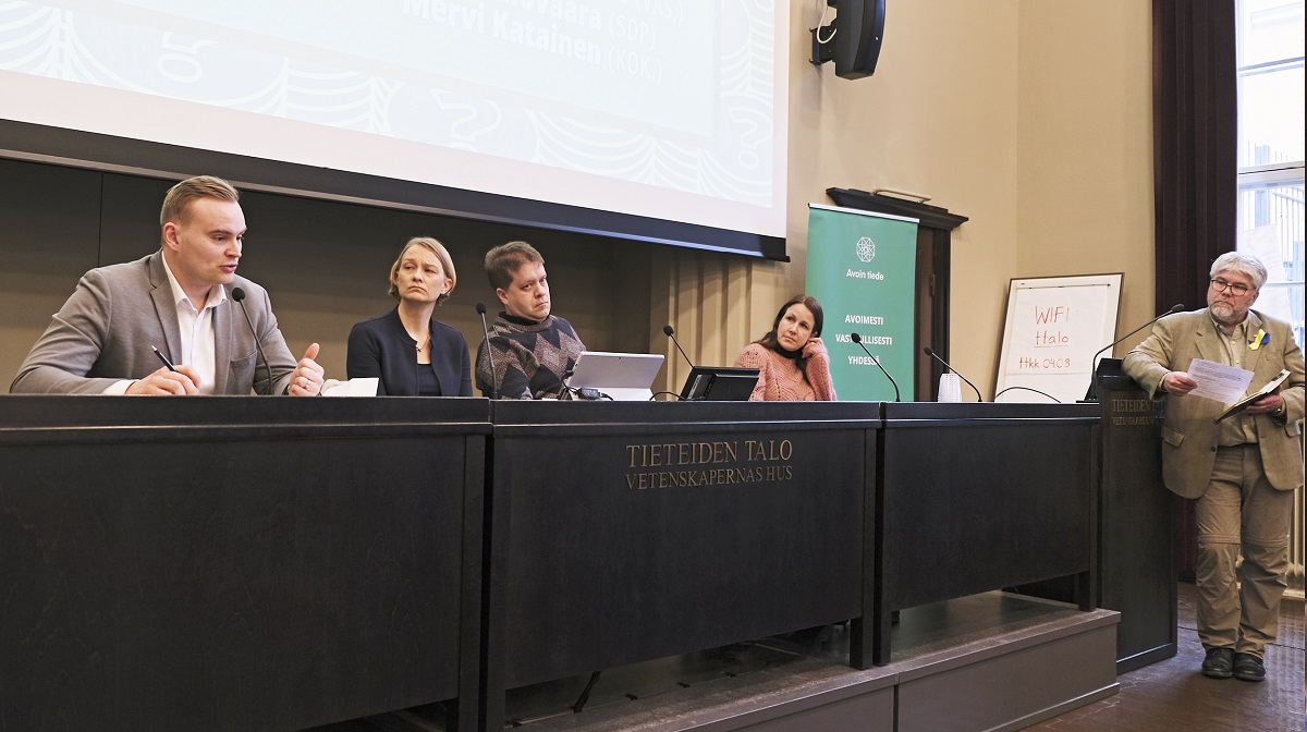 Neljä henkilöä istuu panelisteina ja yksi henkilö seisoo panelistipöydän vieressä kuunnellen panelistin puhetta.