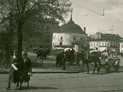 Människor och hästvagnar på torget. En rund, vit byggnad syns i bakgrunden.