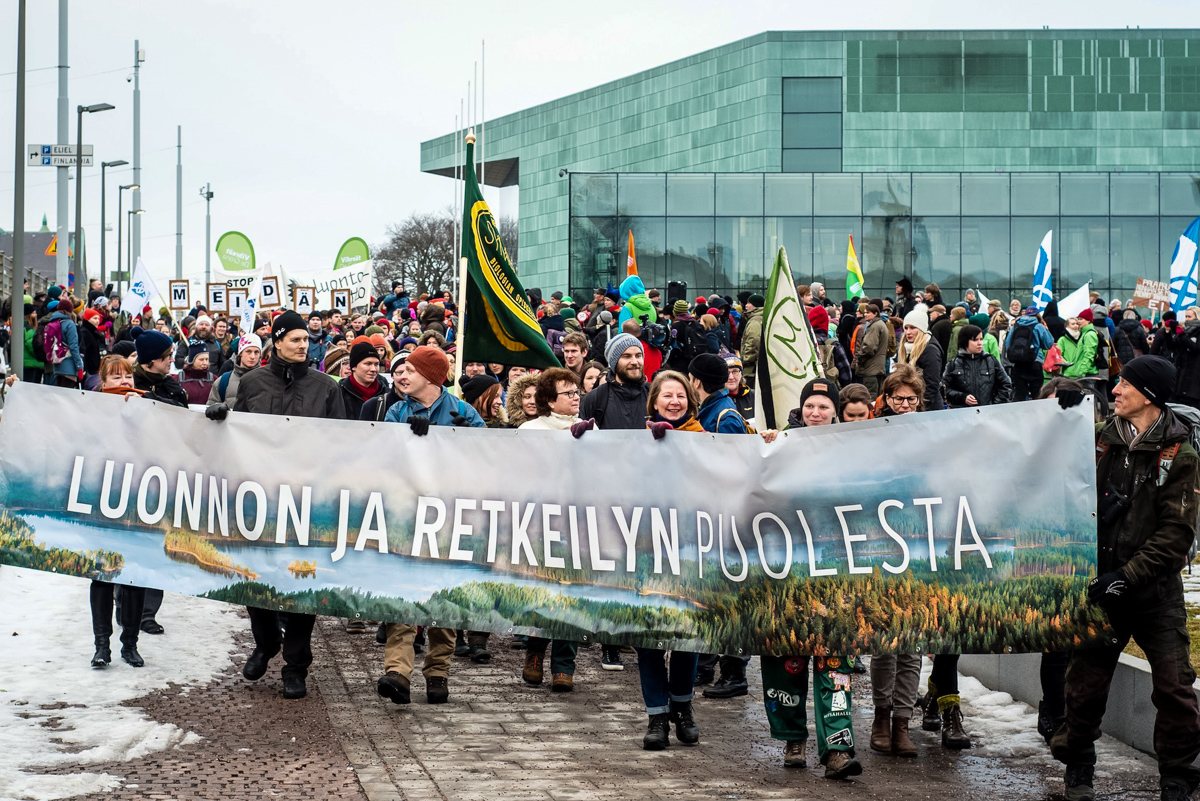 Luonnonsuojeluliiton mainoslakanassa teksti "Luonnon ja retkeilyn puolesta", taustalla ihmisiä marssimassa ilmaston hyväksi..