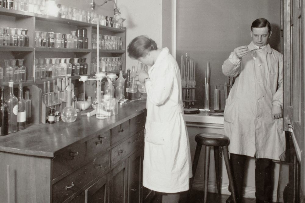 Kaksi henkilöä laboratoriotakeissa tutkimassa näytteitä, taustalla erilaisia lasipulloja ja mittalaitteita.