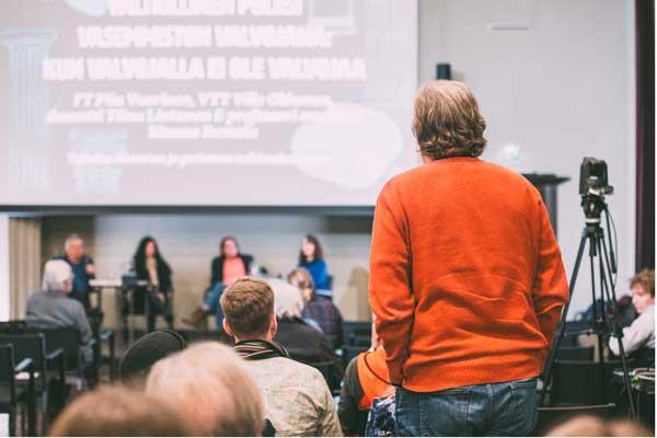 Henkilö punaisessa paidassa on noussut seisomaan luentosalissa esittääkseen kysymyksen. Hänen edessään näkyy lavalla istuvia panelisteja.