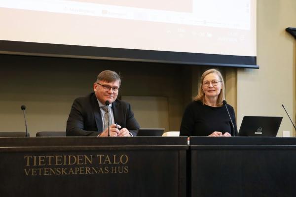 Kaksi henkilöä istuvat puhujanpöydän vieressä ja katsovat kameraan, taustalla näkyy valkokangas.