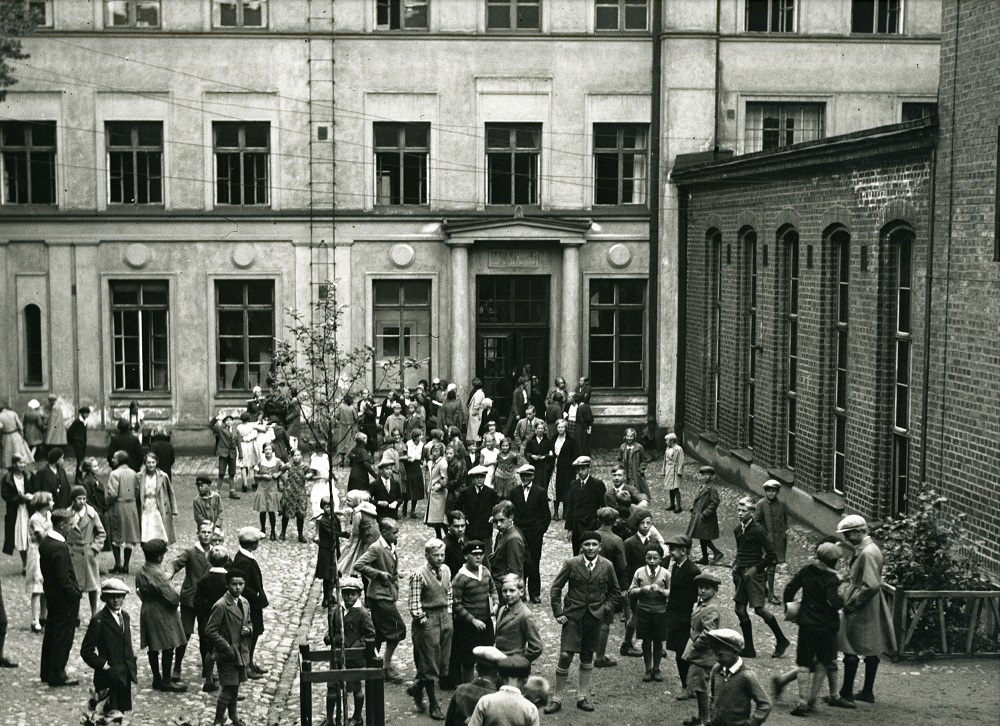 Historiallinen mustavalkoinen kuva, lapsia leikkimässä koulun sisäpihalla.